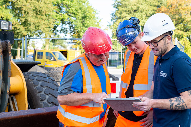 Drei Bauarbeiter in Bauhelmen und Warnwesten betrachten Baupläne auf einem Tablet