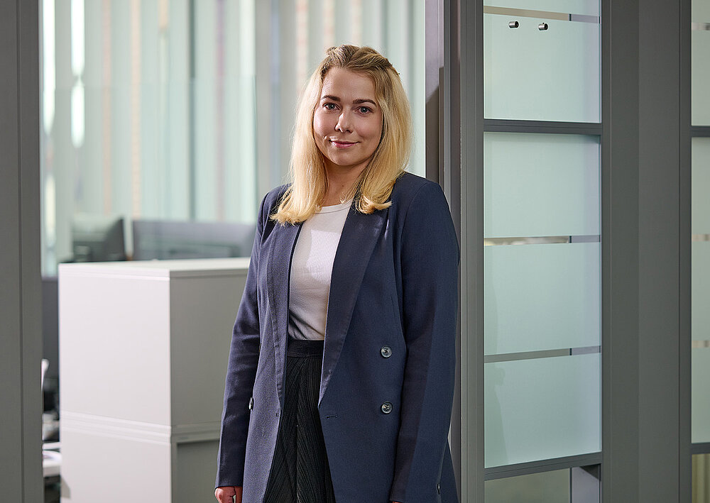 Portrait einer lächelnden jungen Frau in dunklem Blazer vor gläserner Bürotür