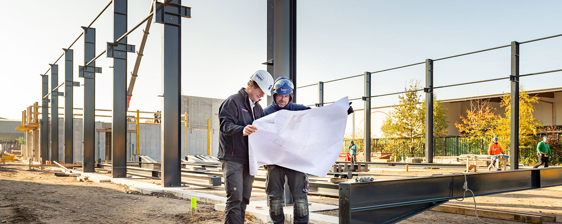 Zwei Bauarbeiter in Schutzkleidung betrachten Baupläne auf einer Baustelle mit Stahlträgern