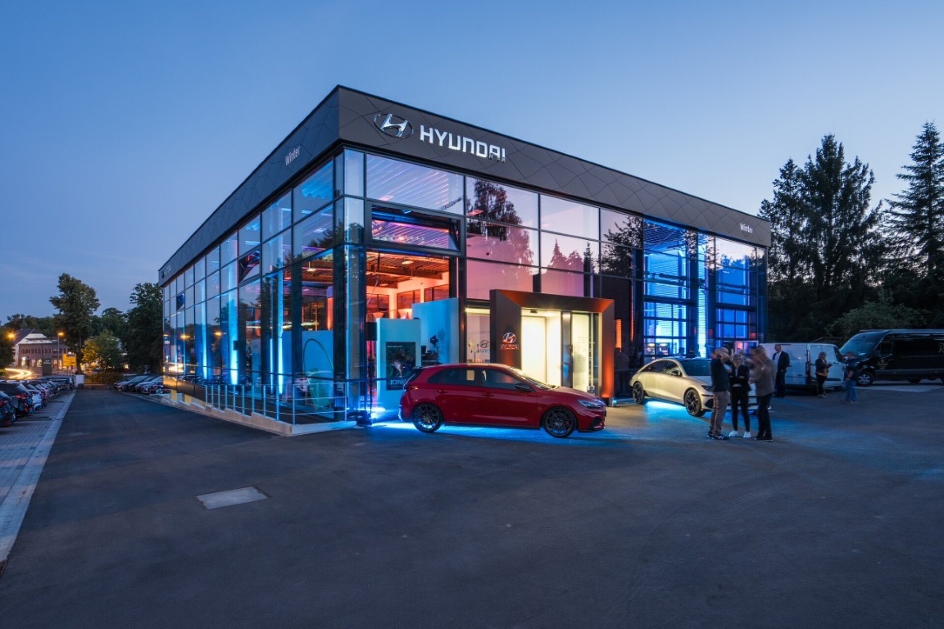Abendliche Außenansicht eines beleuchteten Autohauses mit Glasfassade und einem roten Sportwagen